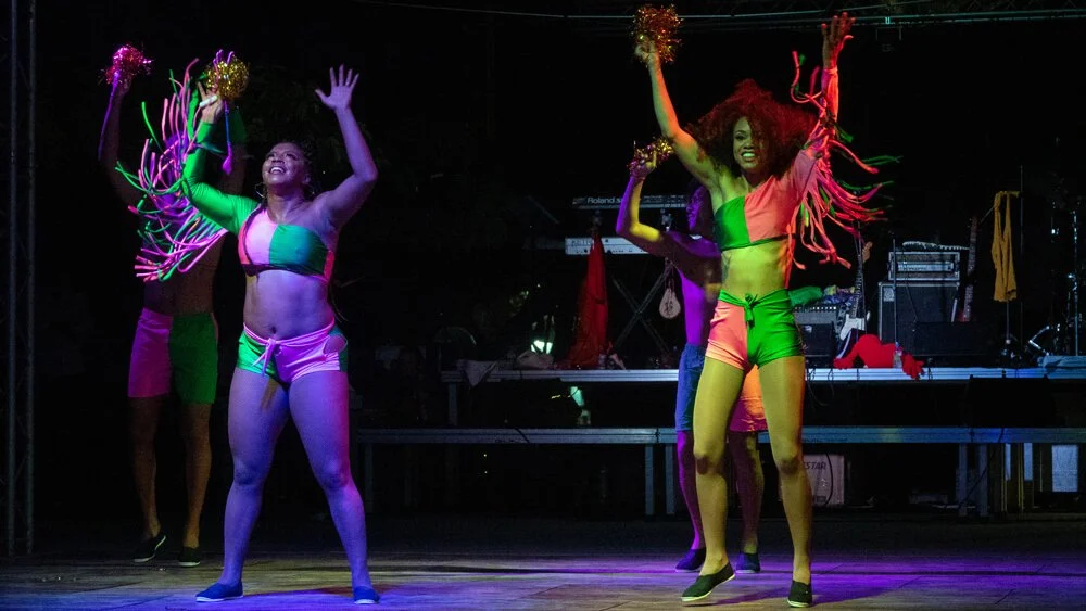 Personas bailando en un escenario con luces de neón y fondo oscuro, usando ropa colorida y accesorios brillantes.