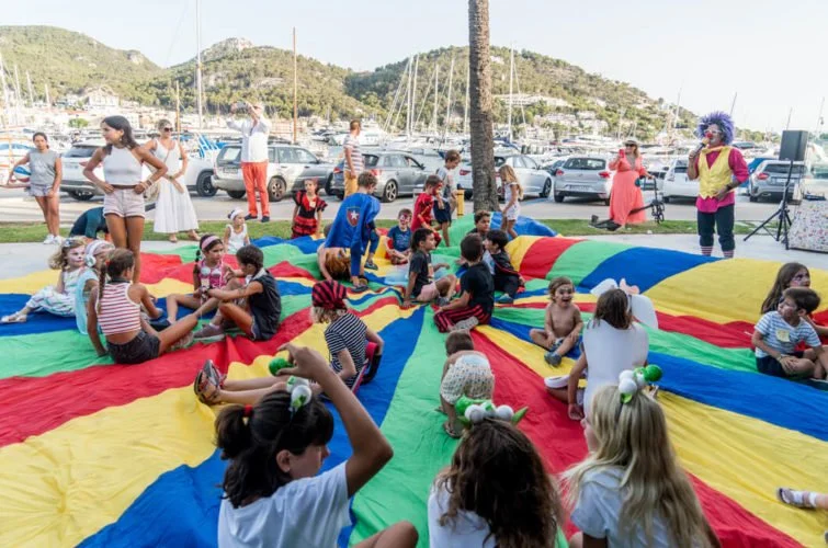Niños jugando en un paracaídas multicolor en un evento al aire libre cerca del puerto, con adultos y un payaso en un fondo de montañas y autos.