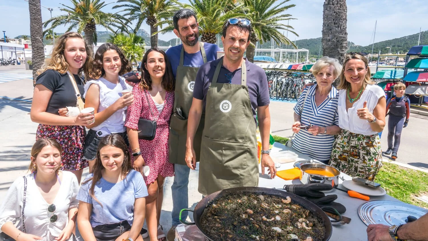 Grupo de personas en una feria de comida al aire libre con algunos platos y una paellera en primer plano, árboles y barcos en el fondo.