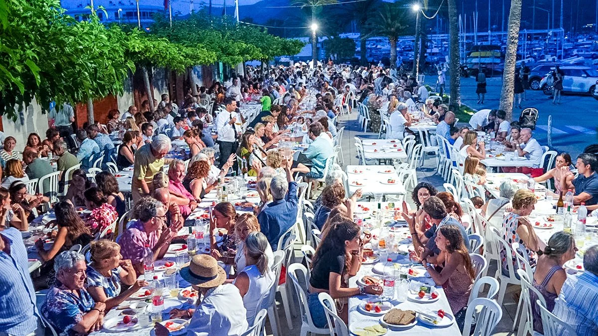 Gran cena al aire libre con muchas personas sentadas en mesas en una calle, parece ser una celebración nocturna.