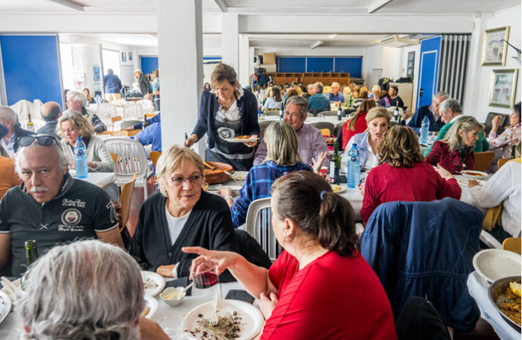 varios adultos comiendo en un comedor con mesas y sillas, algunos en conversación y otros sirviéndose comida