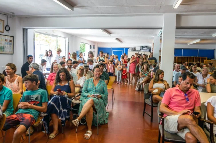 Grupo de personas sentadas y de pie en una sala, algunas usando smartphones, participando en un evento social o reunión.