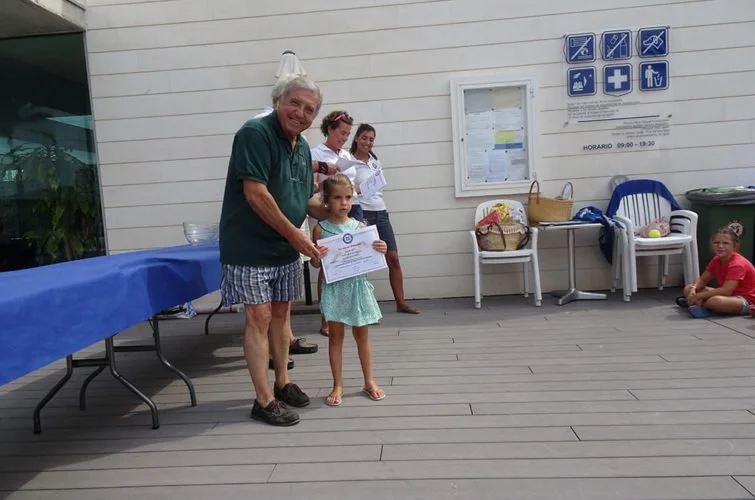 Un hombre mayor entregando un certificado a una niña en un evento al aire libre.