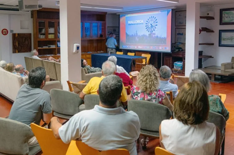 Personas sentadas en una sala viendo una presentación en una pantalla que dice "Mallorca" con un fondo de un parque de diversión.