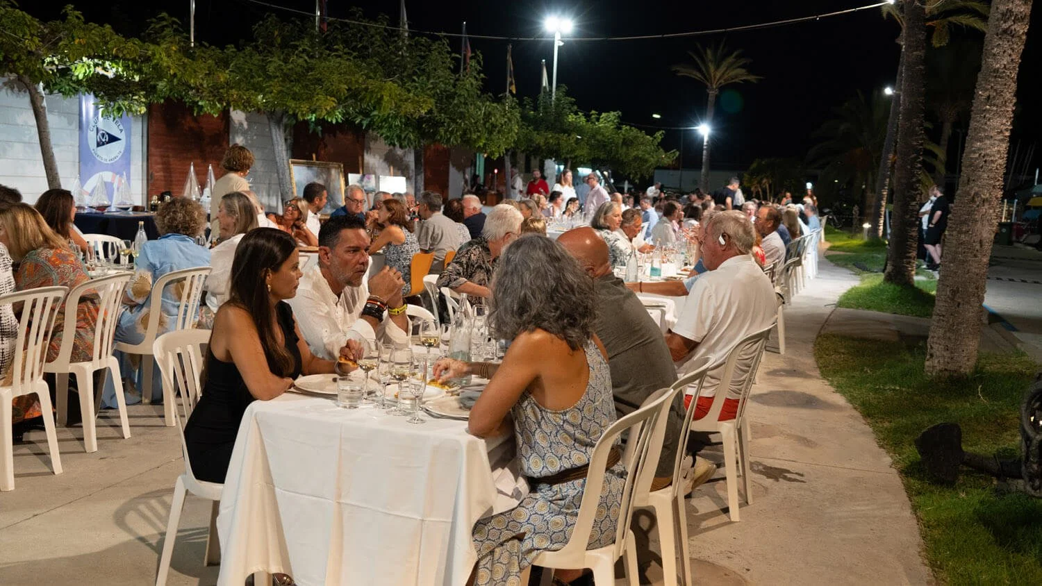Personas sentadas en mesas blancas en un evento al aire libre por la noche, con árboles y luces en el fondo.