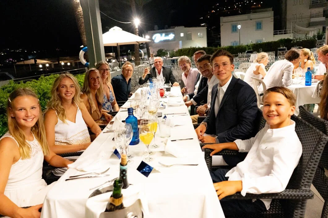 Grupo de personas en una cena al aire libre en la noche, sentados alrededor de una mesa con alimentos y bebidas.