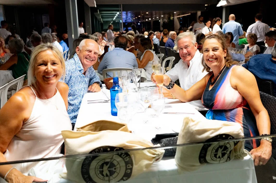 Grupo de cinco personas sonriendo y brindando en una cena en un restaurante elegante, con muchas personas más al fondo.
