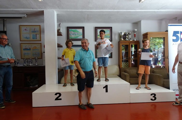 Niños en el podio de una competencia infantil, celebrando en una ceremonia de premiación. Un adulto en el centro posa frente a ellos, mientras otros adultos observes en el fondo.