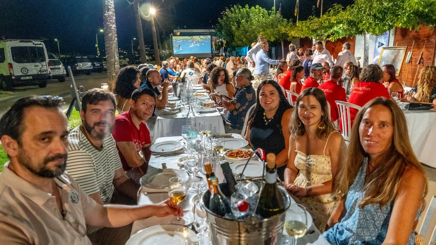 Grupo de personas cenando en una cena al aire libre por la noche, con mesas largas, platos, copas de vino y una pantalla de televisión en el fondo.