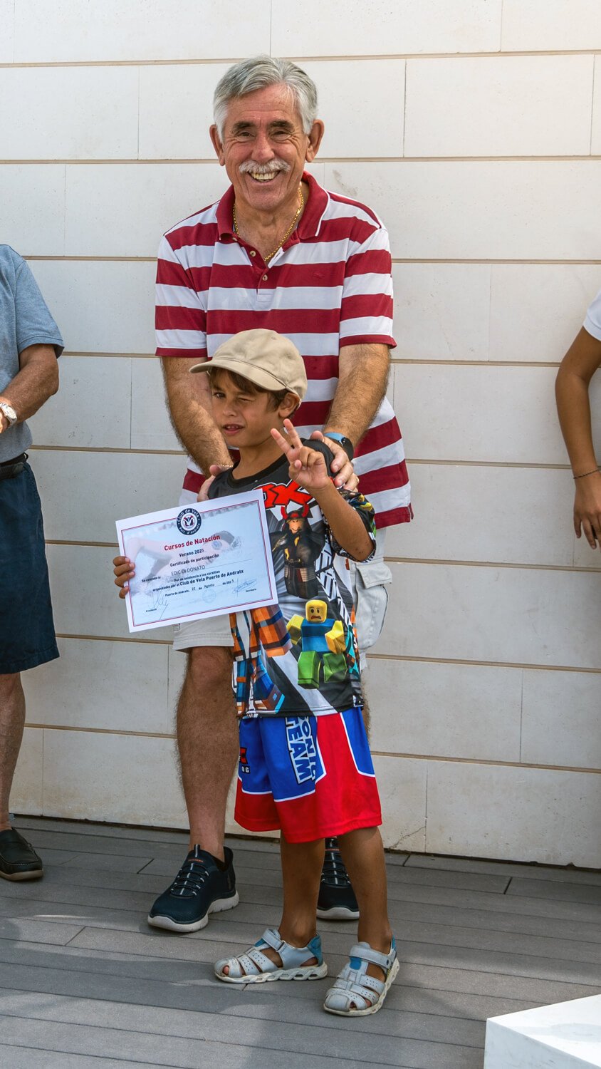 Un niño sosteniendo un diploma de participación en un evento, posando con un hombre mayor que está detrás de él, ambos sonriendo y haciendo una señal de paz con la mano. La ceremonia parece ser relacionada con cursos de natación.