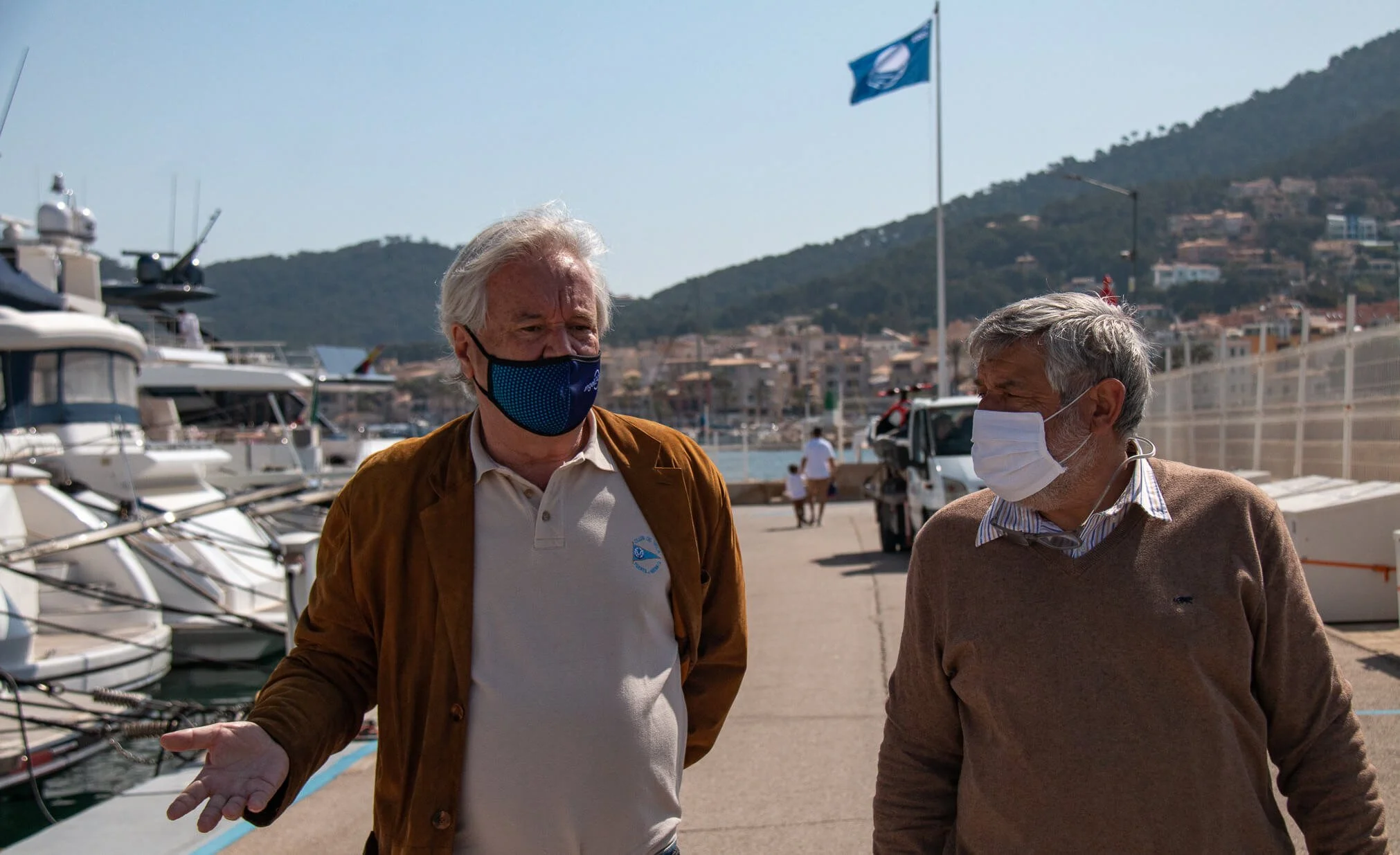 Dos hombres mayores con mascarillas conversando en un puerto con yates y barcos en el fondo, montañas y banderas en el cielo