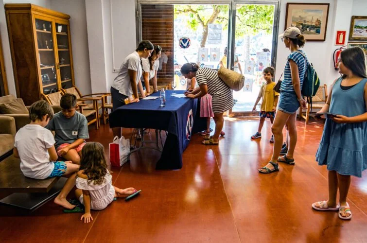Varias personas, tanto adultos como niños, esperan en una sala de interior con suelo de color marrón. Algunos niños están sentados en el suelo, otros en una banca y algunos adultos en línea en una mesa con una cubierta azul. La entrada es de vidrio y