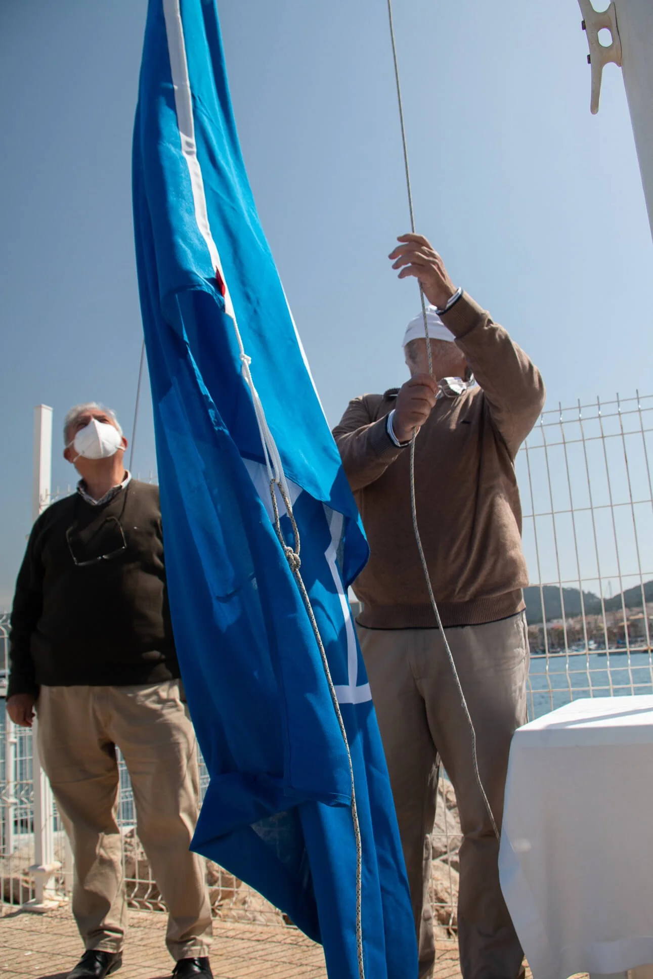 Dos hombres con mascarillas ajustan una bandera azul en un muelle, con una cerca de fondo y un cielo despejado.