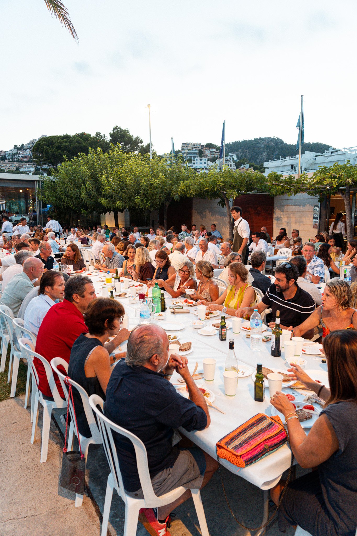 Grupo de personas disfrutando de una comida en una mesa larga en un evento al aire libre durante la tarde o noche, con árboles y edificaciones en el fondo.