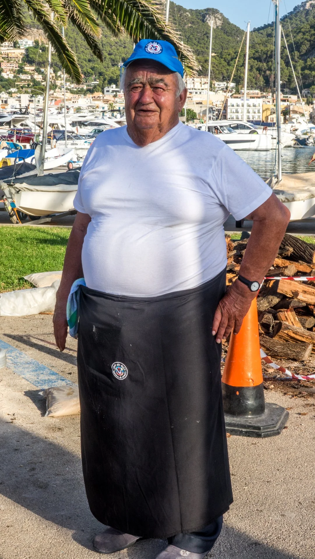 Un hombre viejo con camisa blanca, falda negra y gorra azul en un puerto de yates, con montañas y edificios en el fondo.