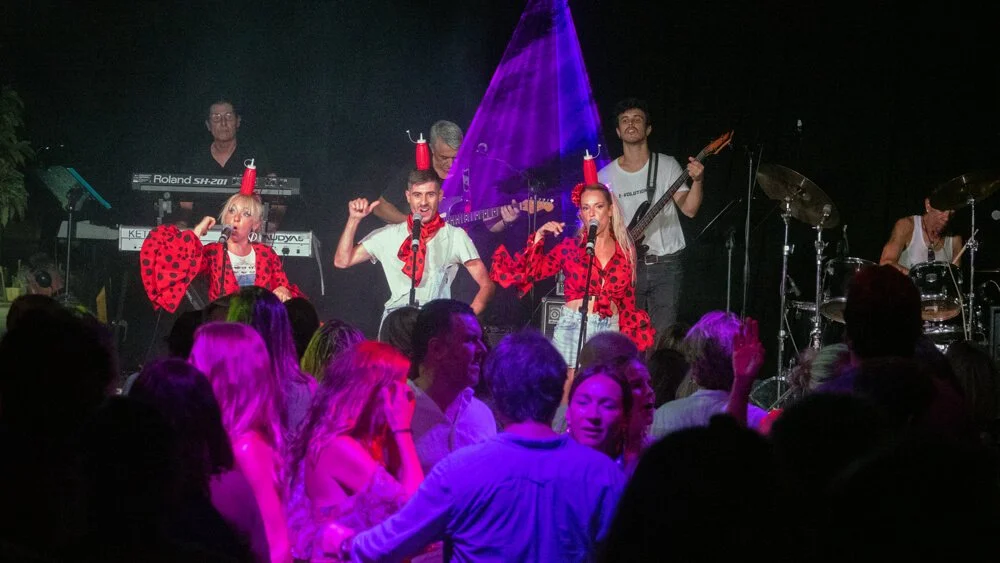 Conjunto musical en un concierto, con miembros usando ropa con estampados de lunares rojos y negros, tocando instrumentos y cantando frente a una audiencia. La iluminación es colorida y el ambiente es festivo.