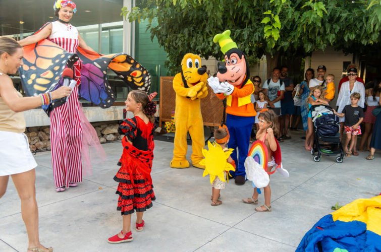 Niños en un espectáculo con personajes de dibujos animados y un payaso, en un espacio al aire libre, con adultos observando.