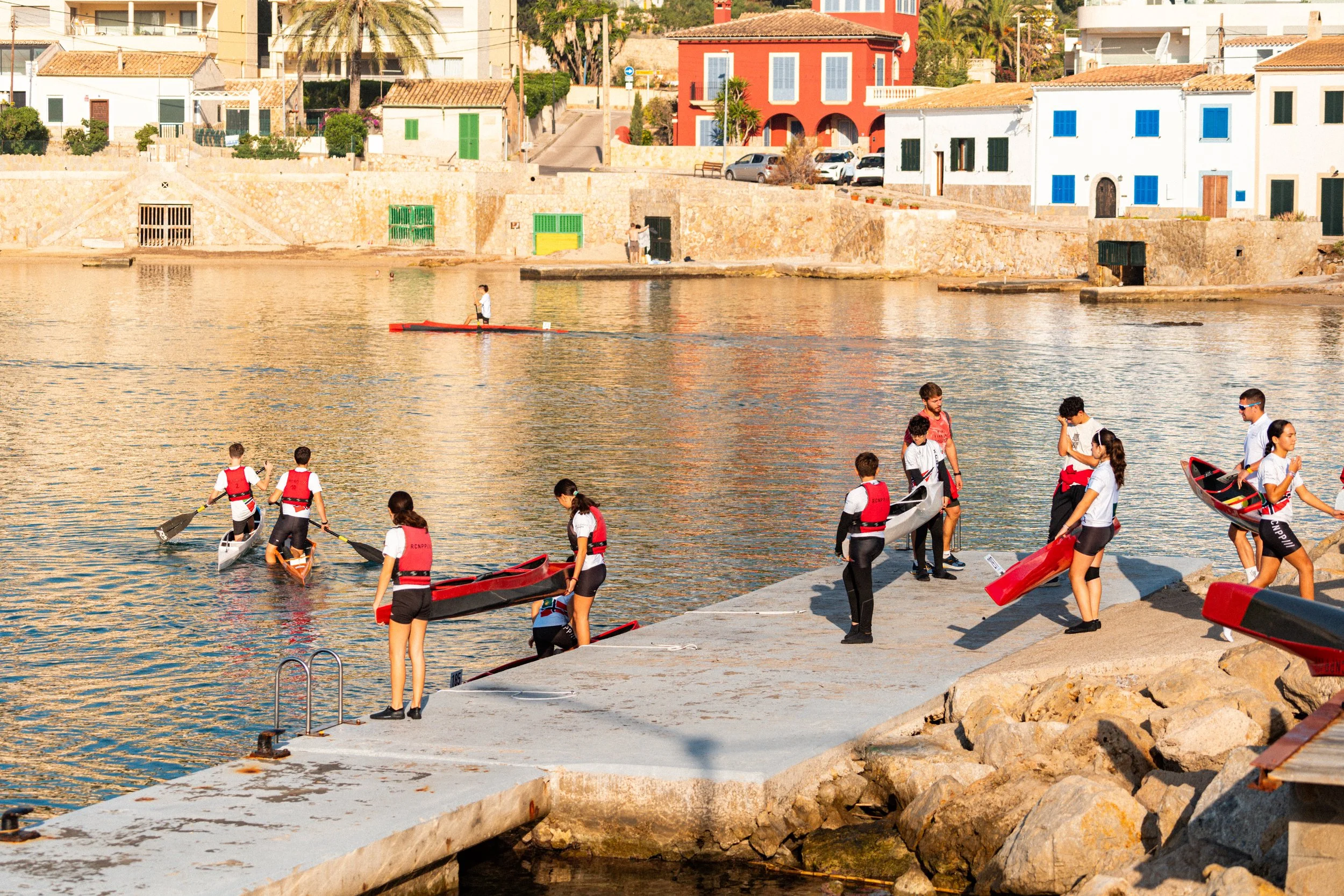 Un grupo de niños y adolescentes con chalecos salvavidas disponiéndose a participar en actividades acuáticas en un muelle junto a un río o lago, con casas coloridas y edificios en el fondo.