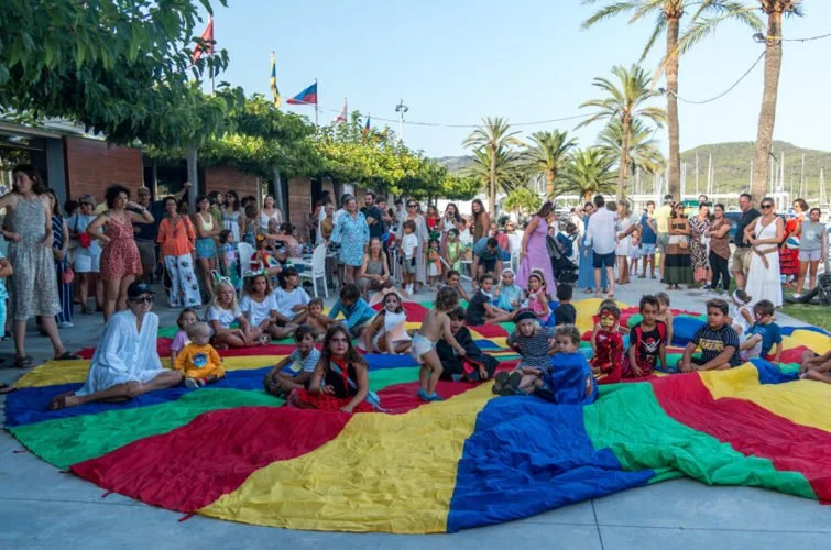 Niños y adultos participando en un juego con un paracaídas colorido en un espacio exterior con palmeras, árboles y banderas