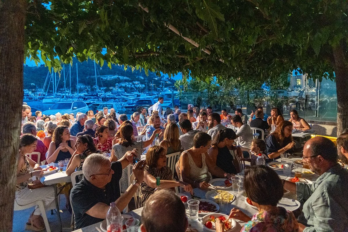 Cena de una reunión social al aire libre en la noche con muchas personas sentadas en mesas comiendo y bebiendo, bajo árboles y con vista a un puerto con yates y barcos.
