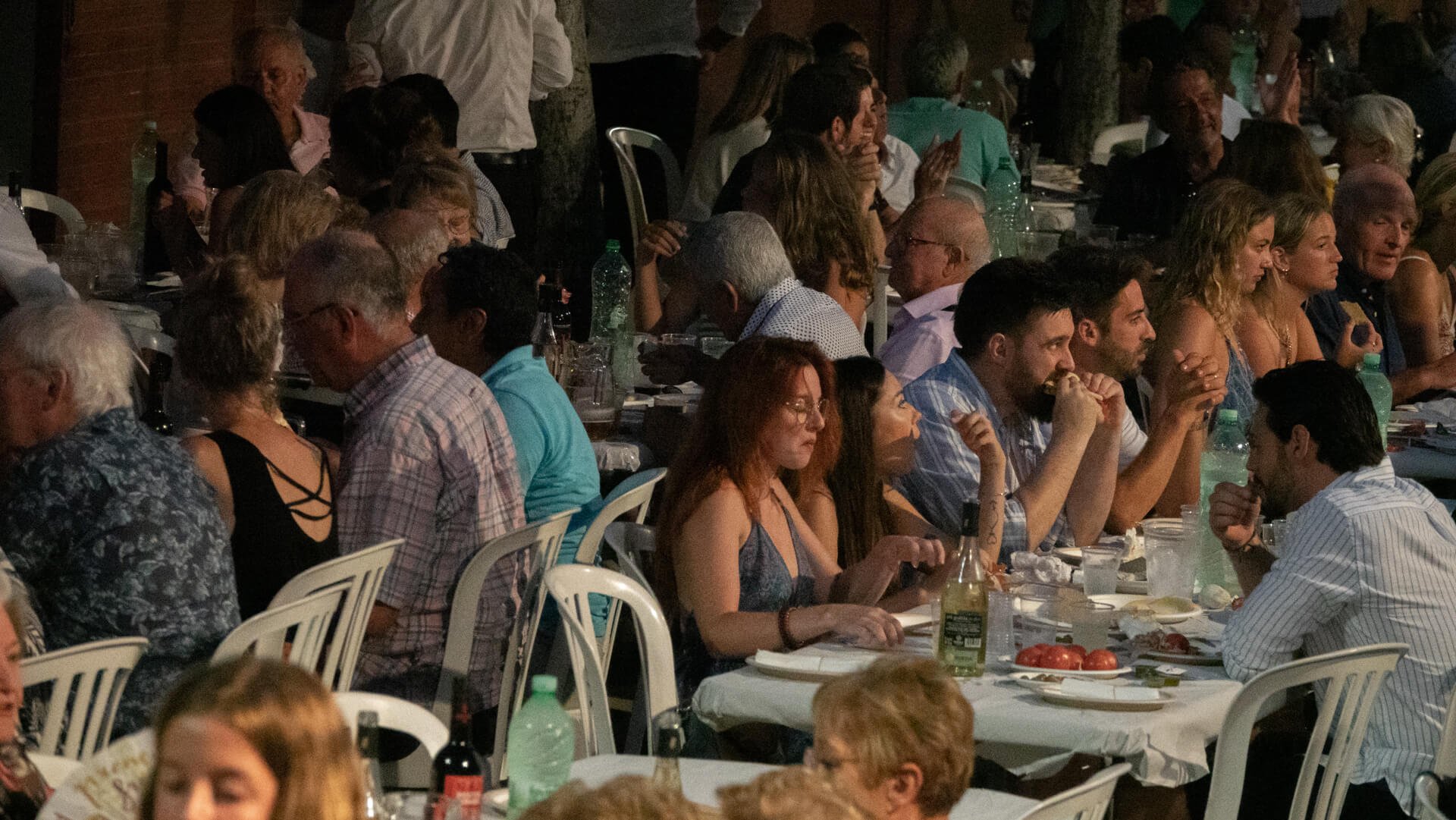 Un grupo de personas sentadas en mesas largas en una cena o evento social en un ambiente nocturno. La mesa tiene botellas de agua, vino y comida. La iluminación es tenue y la escena muestra a varios individuos conversando y disfrutando.