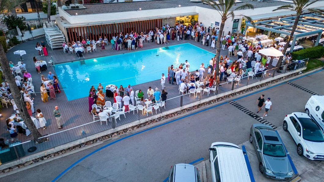 Celebração ao lado de uma piscina com muitas pessoas, mesas e cadeiras, em um espaço ao ar livre com palmeiras e carros na frente.