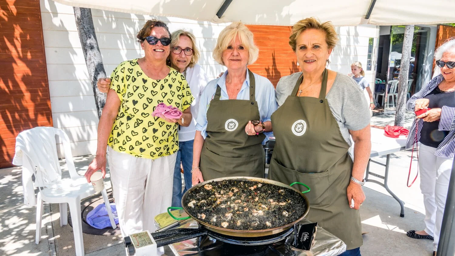 Grupo de mujeres mayores cocinando en una sartén grande al aire libre, con algunas sonriendo y otras observando, en un entorno exterior con sombra y mesas.