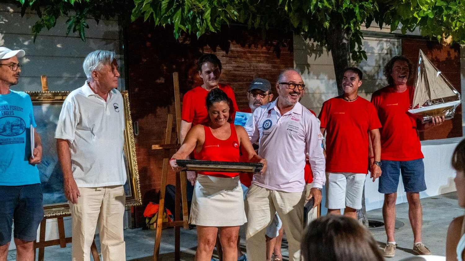 Grupo de personas en una ceremonia al aire libre, algunas sosteniendo premios y objetos, en un ambiente festivo nocturno con árboles y decoraciones.
