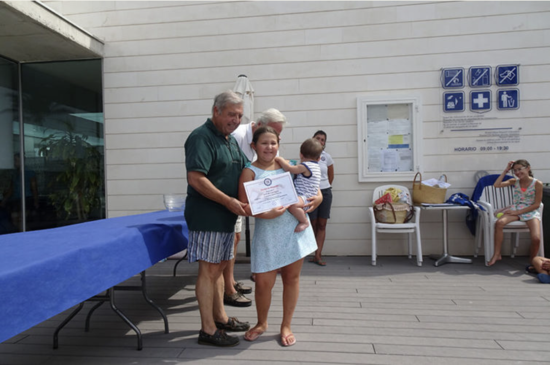 Una mujer sostiene un certificado y un bebé en brazos, mientras un hombre mayor le entrega un reconocimiento en una ceremonia al aire libre. En el fondo, otras personas observan y la pared de un edificio muestra señales y avisos.