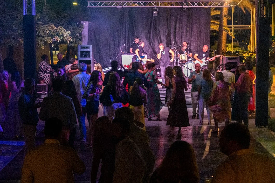 Escenario con banda tocando en vivo y personas bailando en un espacio abierto iluminado con luces de colores, en un entorno nocturno.