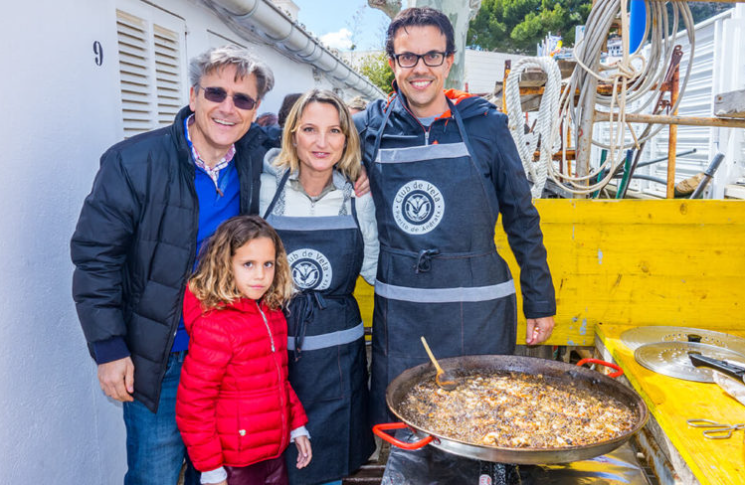Cuatro personas sonrientes cocineros y una niña en una calle, con una gran paella en la estufa y un carro cargado de utensilios de cocina.