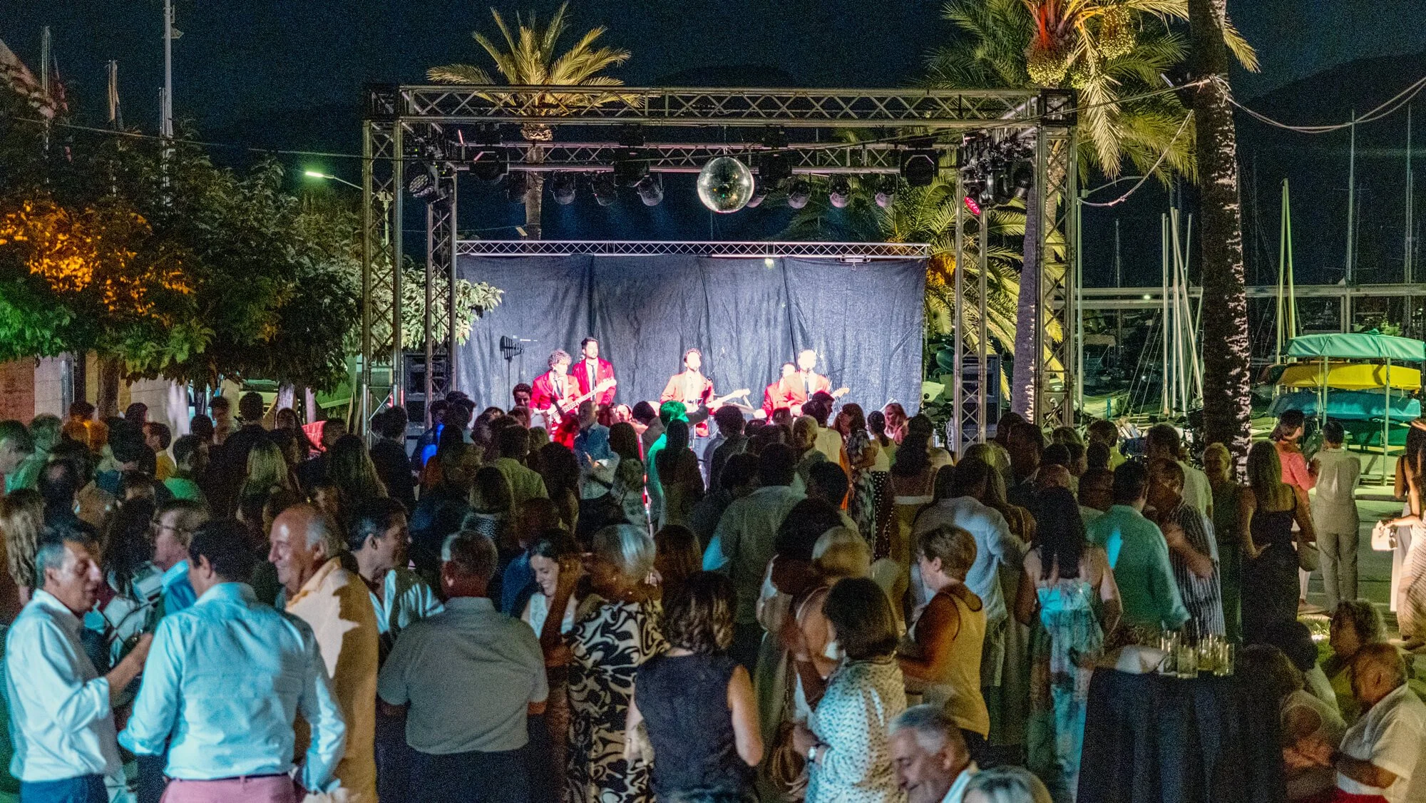 Concierto al aire libre de noche con banda tocando en un escenario y público disfrutando, rodeado de palmeras y vegetación.