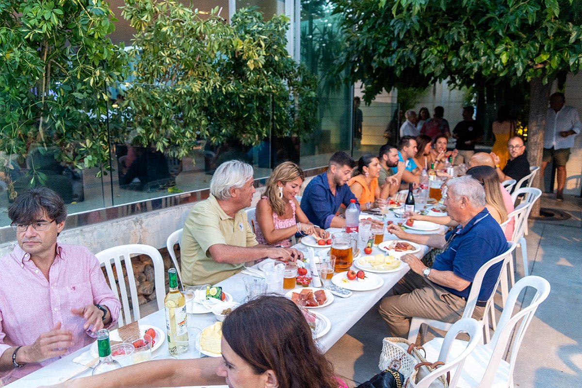 Grupo de personas sentadas en una mesa al aire libre compartiendo comida y bebida durante una reunión social o celebración.