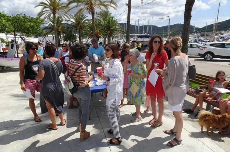 Grupo de personas en un evento al aire libre junto a un puerto con yates, en un día soleado y varias palmas.