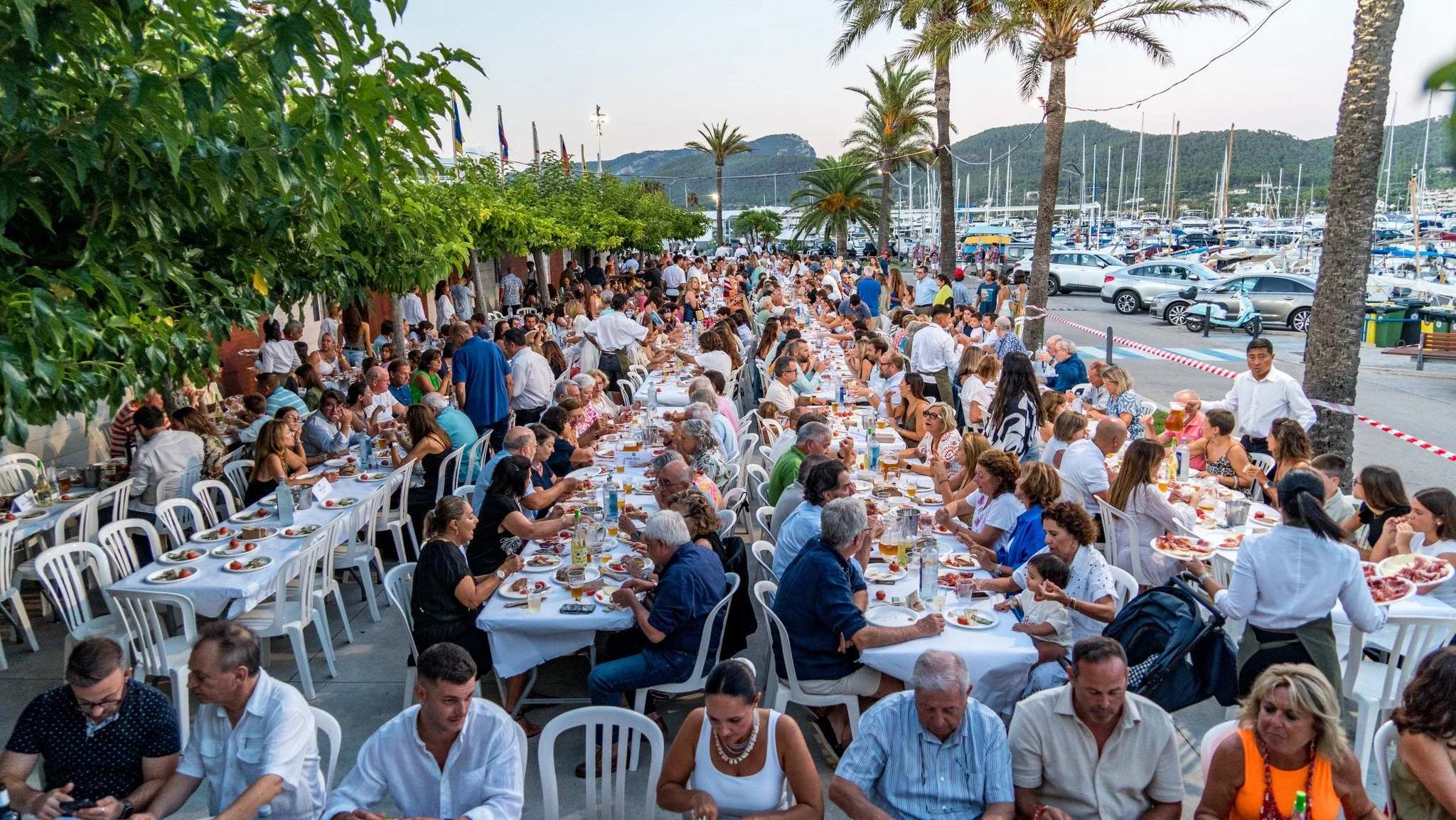 Una multitud de personas celebrando una comida al aire libre junto a un puerto con yates y montañas de fondo, con mesas largas llenas de comida y bebida.