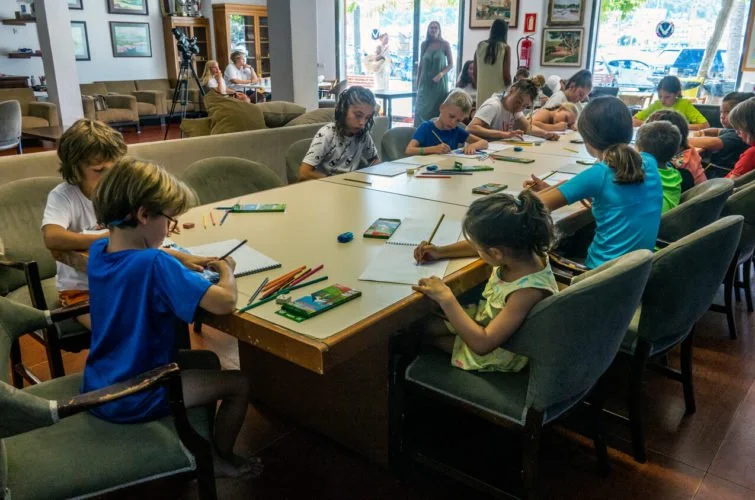 Niños sentados alrededor de una mesa en un salón, dibujando con papeles, lápices y libros de colores, en un espacio interior con ventanas grandes y personas caminando afuera.