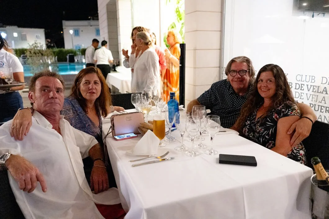 Cuatro personas en una mesa de restaurante con bebidas y utensilios, ambiente elegante y gente en el fondo.