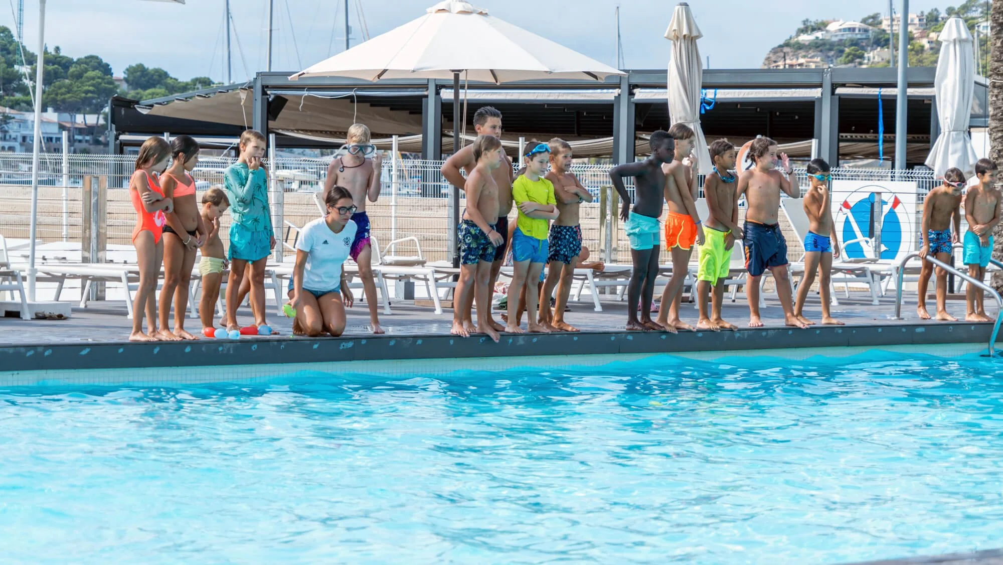 Niños en traje de baño observando en la orilla de una piscina bajo sombrillas en un día soleado
