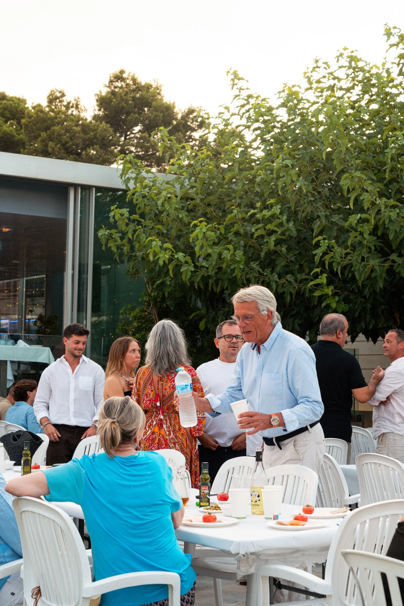 Personas en una reunión social al aire libre, alrededor de mesas con comida y bebidas, algunos conversando, en un entorno con árboles y un edificio moderno.