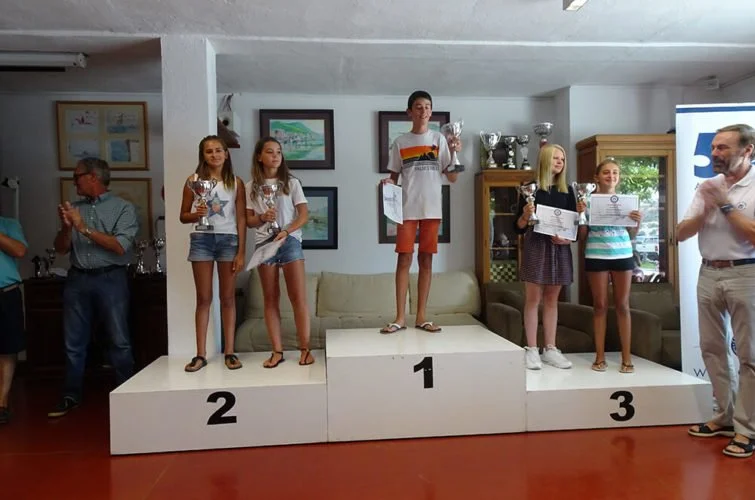 Niños en un podio de premiación, con el primer lugar en el centro y los otros lugares a su izquierda y derecha, sosteniendo trofeos en un ambiente interior.