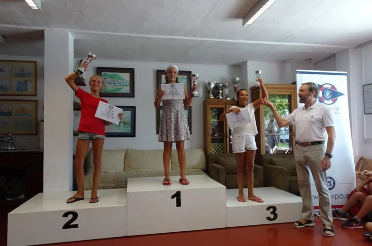Tres niñas en un podio ganando un trofeo, la niña en el primer lugar en el centro, la segunda en la izquierda y la tercera en la derecha, con un hombre entregando el trofeo en una sala con cuadros y ventanas