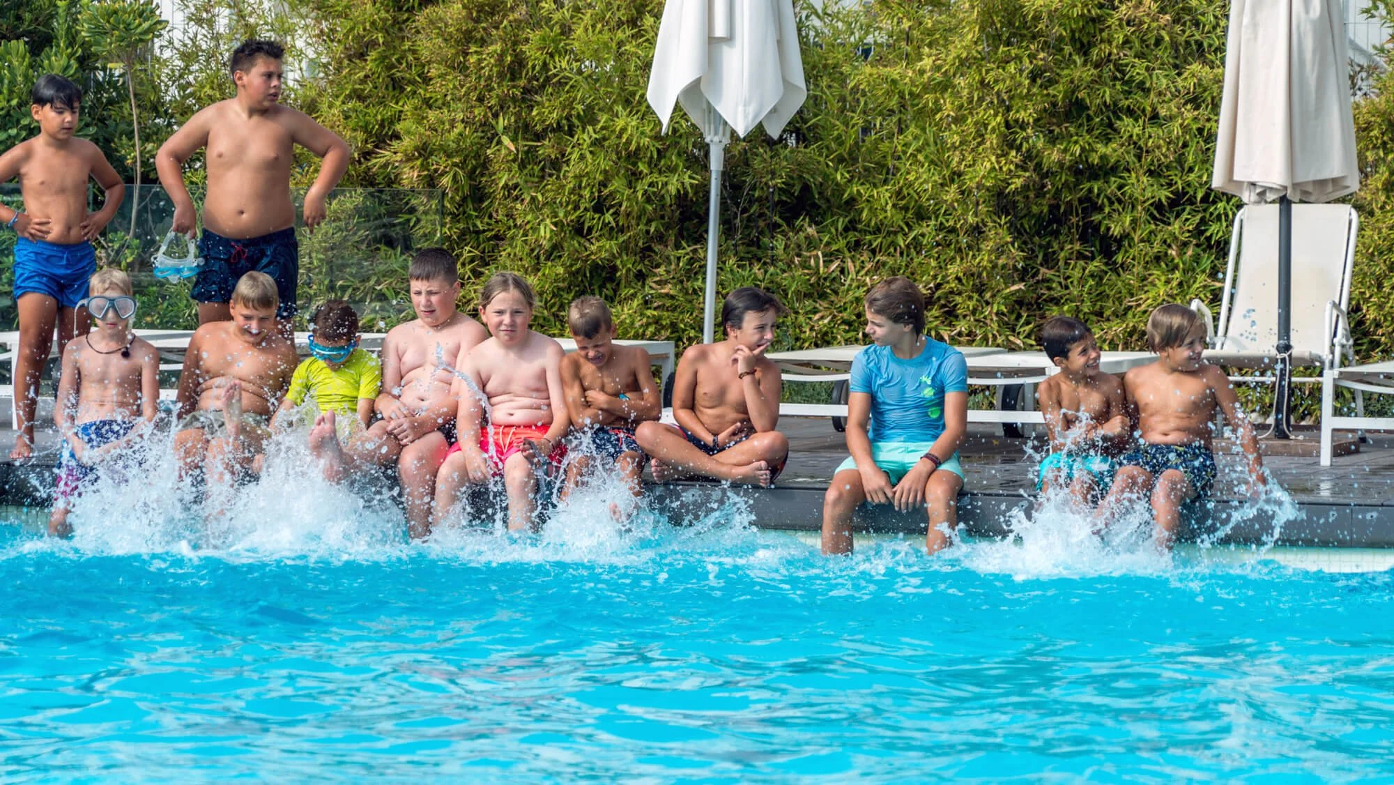 Niños jugando y disfrutando en una alberca al aire libre en un día soleado, algunos están sentados en el borde de la piscina, otros en el agua, con expresiones de diversión y concentración.