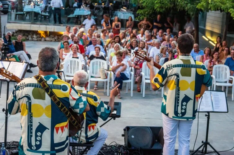 Un grupo de músicos tocando en un escenario al aire libre, con una audiencia sentada frente a ellos, en una especie de patio o plaza.