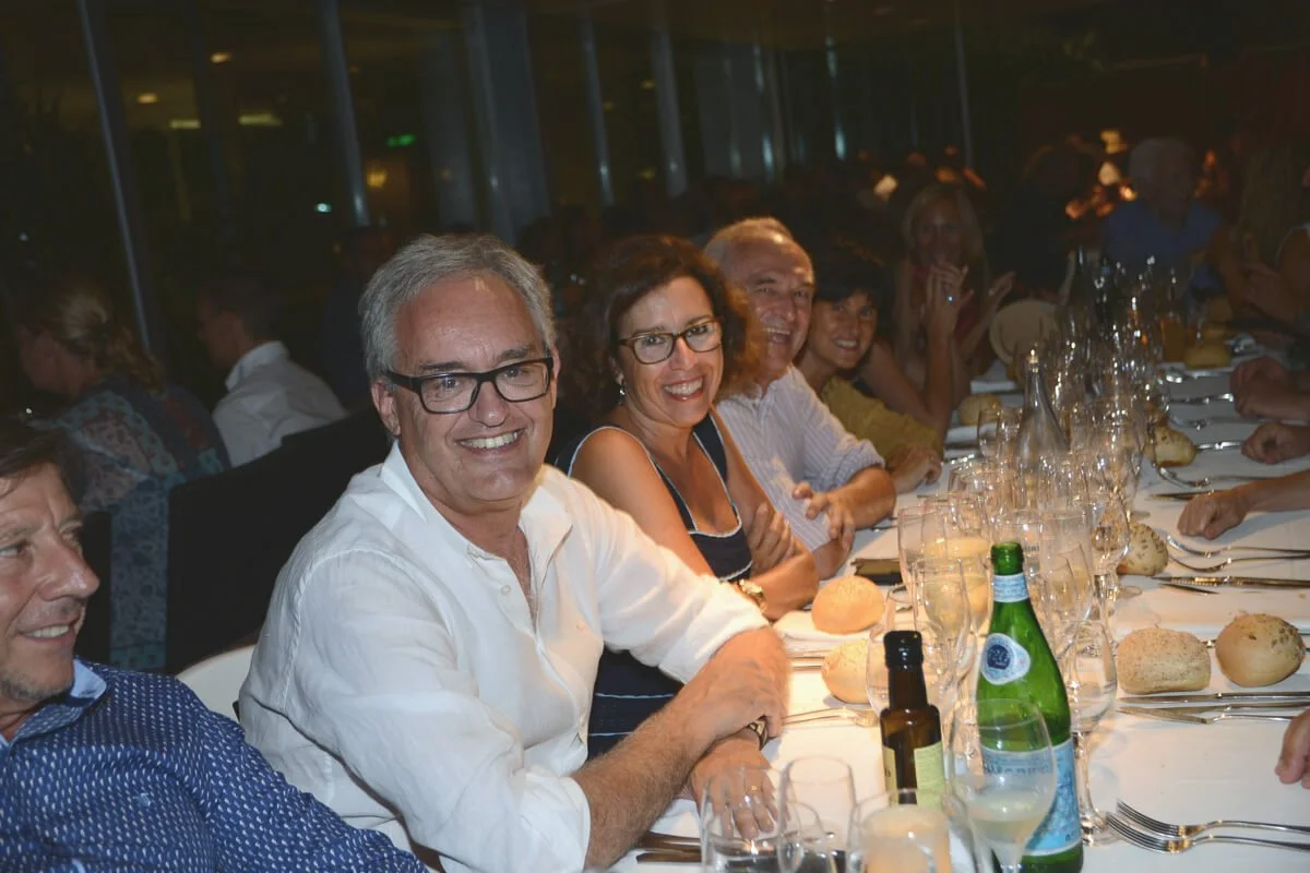 Grupo de personas sonriendo y disfrutando en una cena formal en una mesa larga con panes, botellas de agua y vino, en un ambiente elegante y bien iluminado.