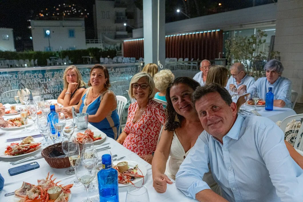 Grupo de personas cenando en una mesa al aire libre en la noche, con comida y bebidas, sonriendo y disfrutando del momento.