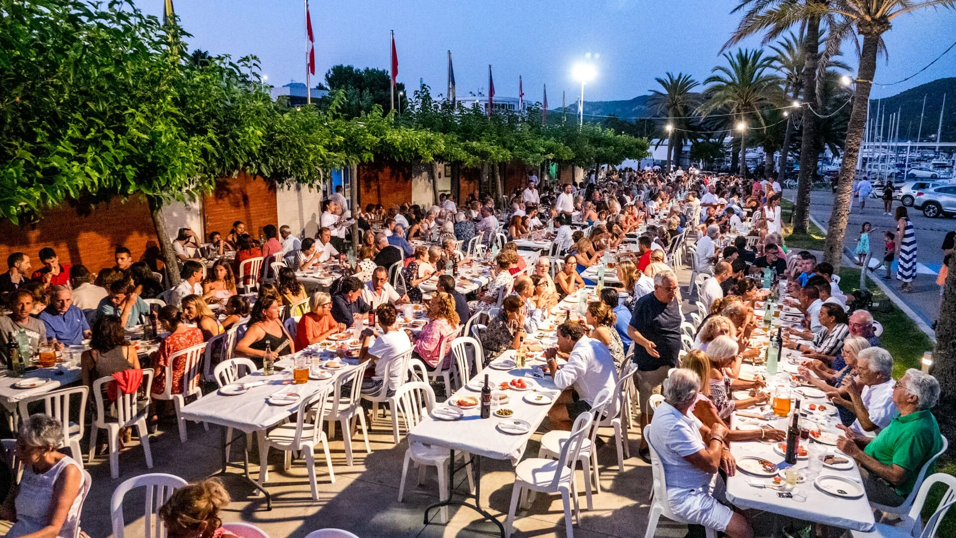 Cena de una cena al aire libre con muchas personas sentadas en mesas largas, comiendo y bebiendo, en un ambiente festivo con muchas luces, árboles y palmeras.