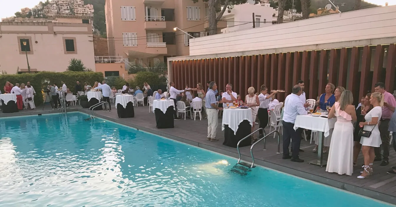 Eventos sociales al aire libre junto a una piscina, con personas conversando y sirviéndose bebidas y comida en mesas con manteles blancos, al atardecer en un espacio urbano.
