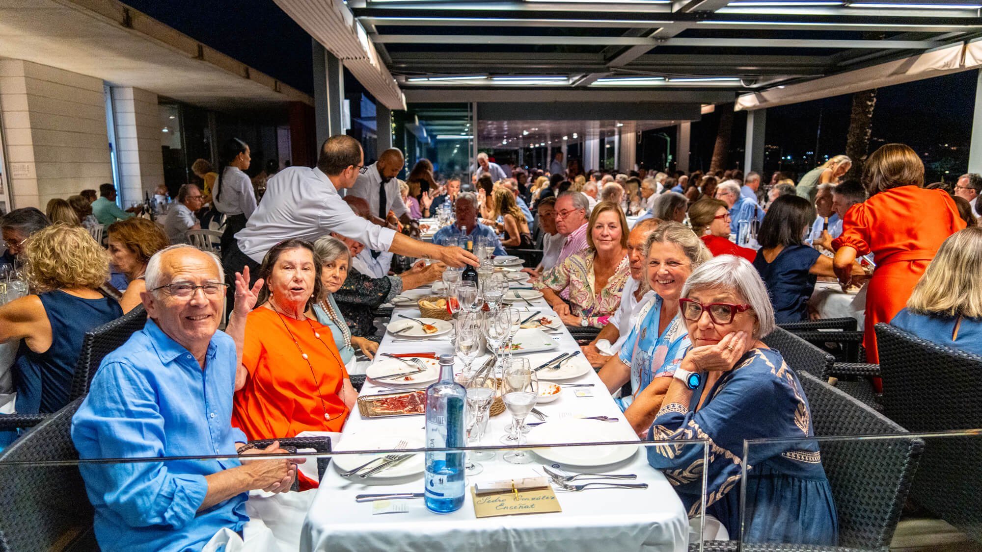 Grupo de personas disfrutando en una cena formal en un restaurante, con mesas largas, comida, bebidas y camareros atendiendo.