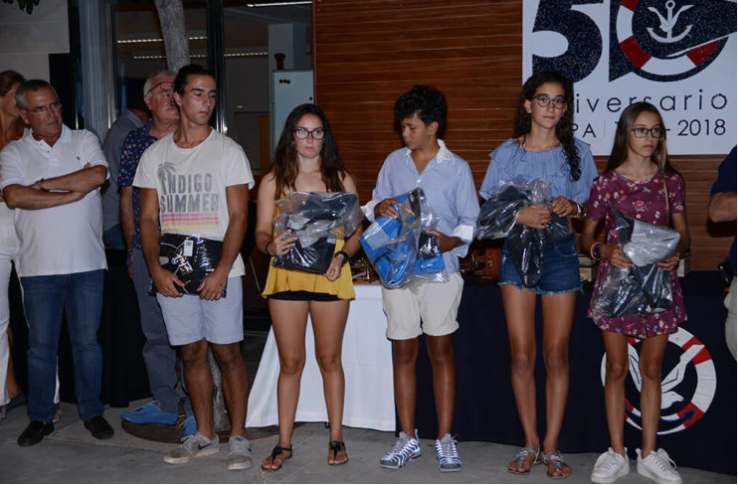 Grupo de jóvenes premiados en una ceremonia con adultos observando, en un evento con un cartel que celebra un aniversario en 2018.