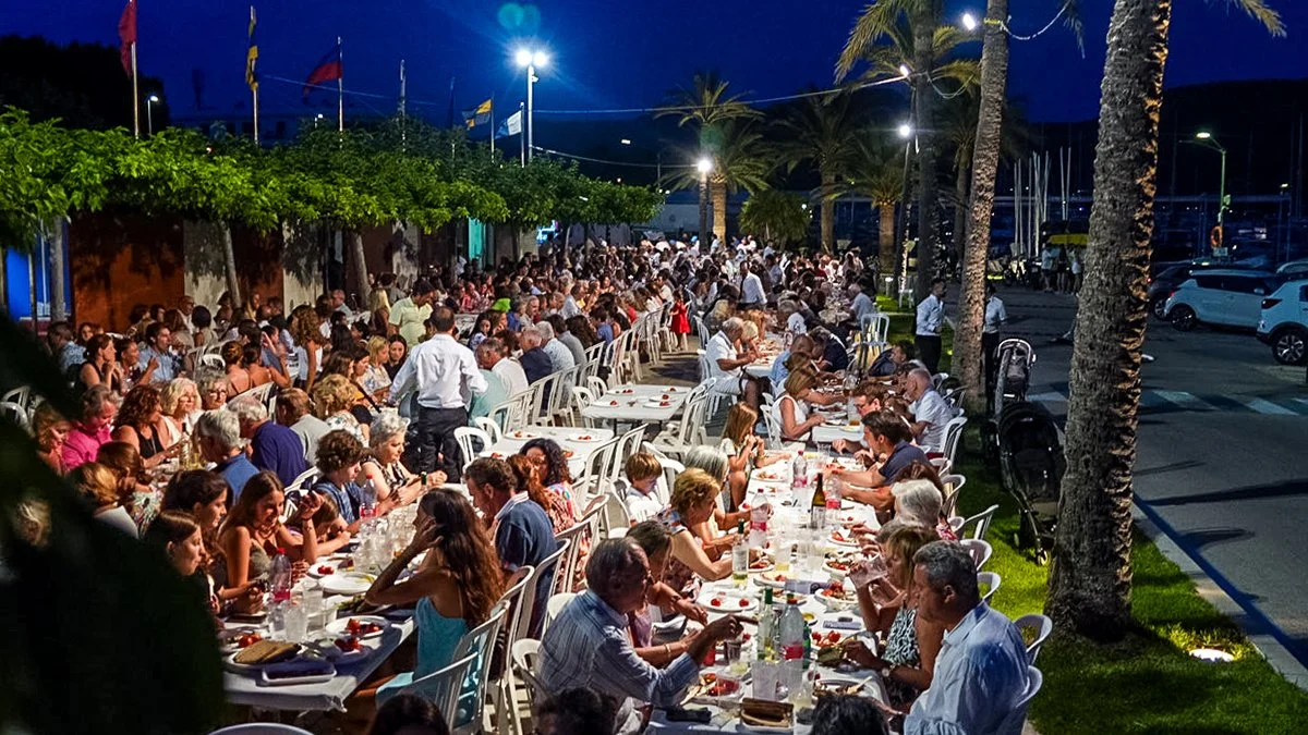 Cena al aire libre en la noche con muchas personas sentadas en mesas, comiendo y socializando, rodeadas de palmeras y luces.